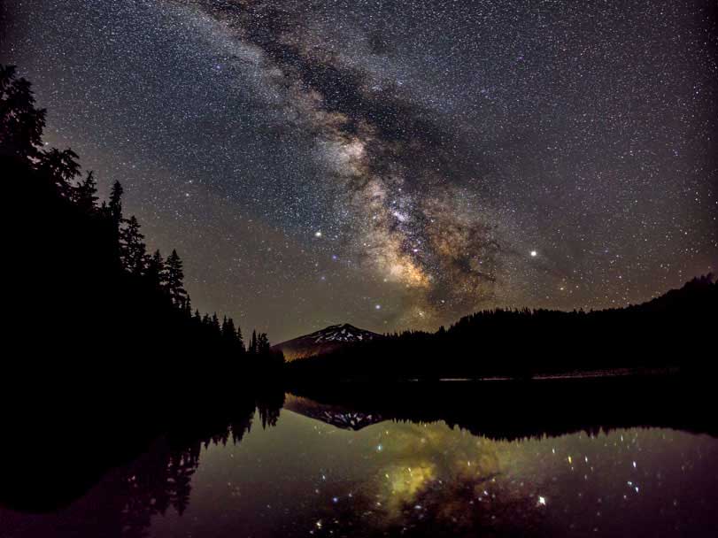 Dark sky showing the milky way, clouds covering the moon, lighting up the surface of Todd Lake, with Mount Bachelor in the distance